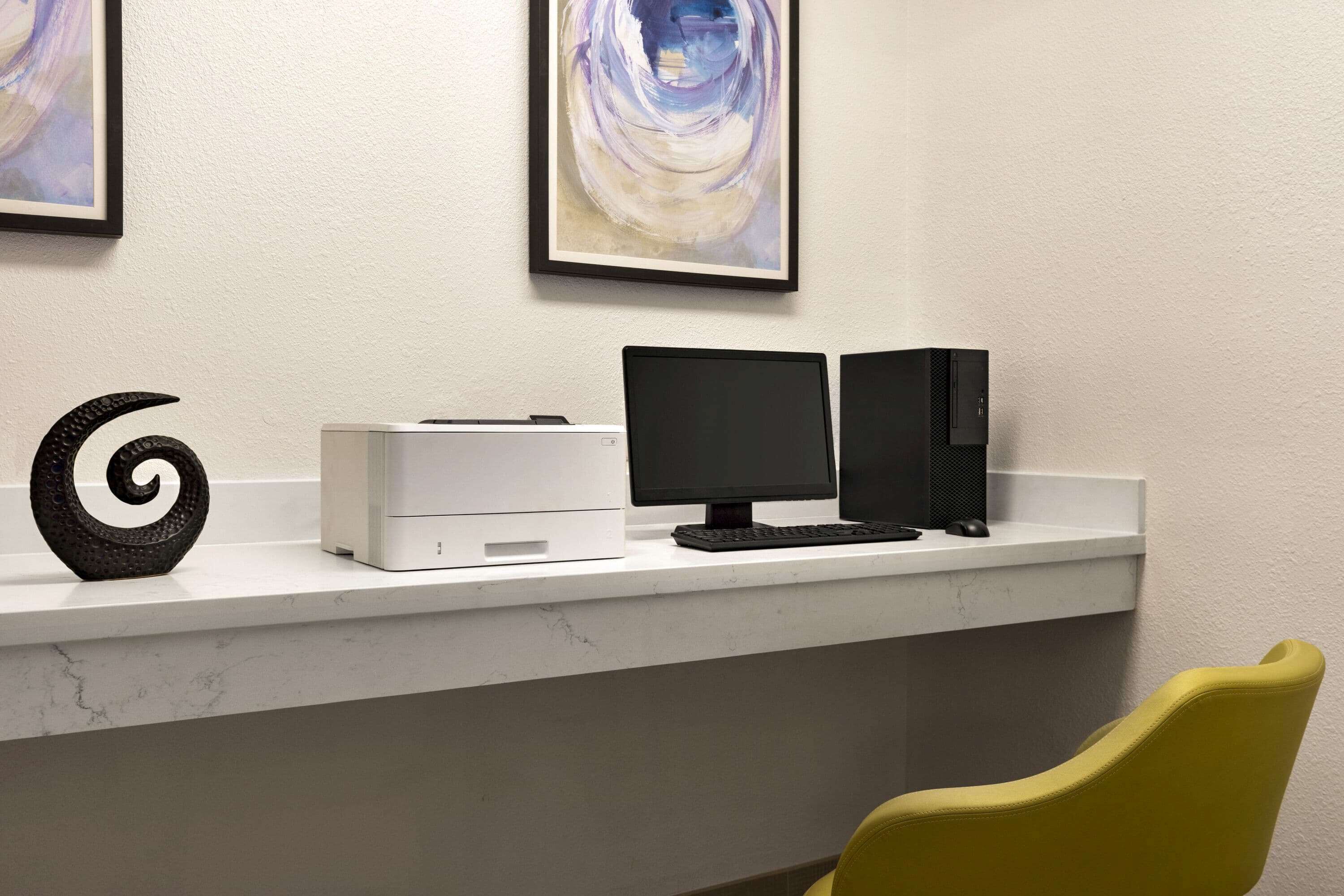A desk with a printer, monitor, and speakers, a spiral sculpture, and a yellow chair.