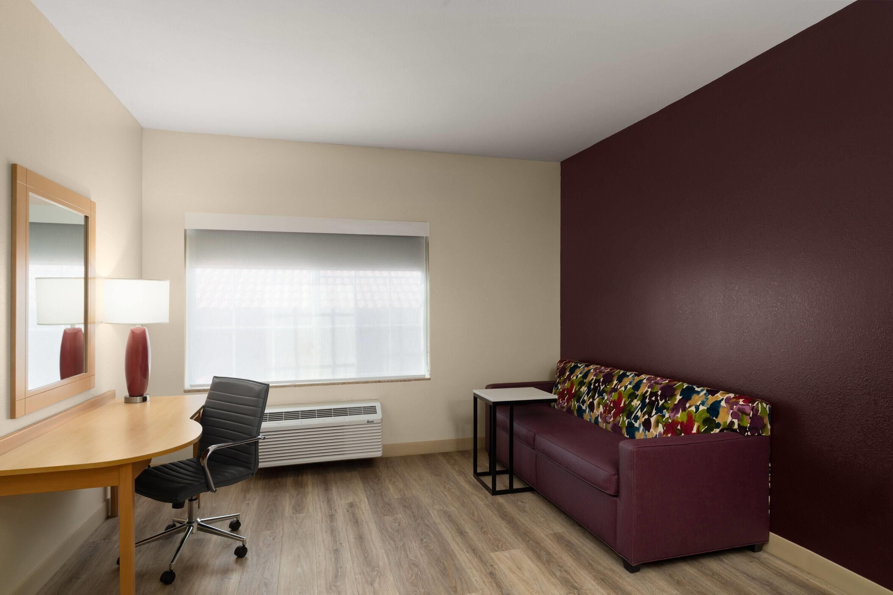 Una habitación con escritorio, silla, sofá y un amplio ventanal con persianas.