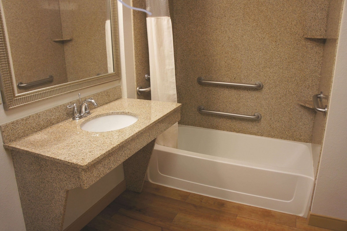 Un baño con lavamanos de encimera de granito, una bañera blanca y una zona de ducha con barras de apoyo.