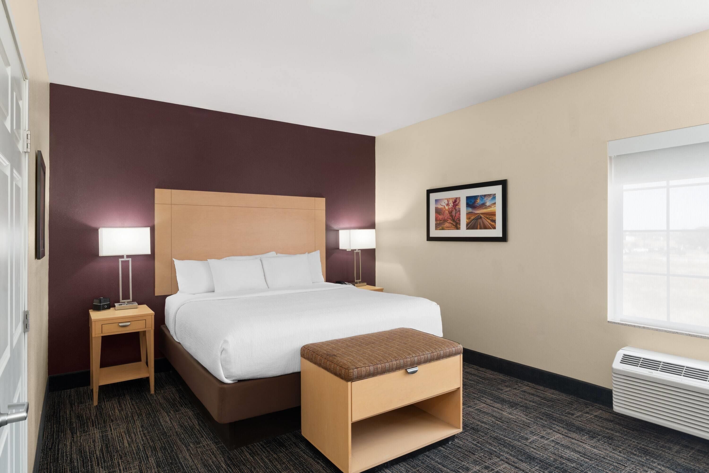 A hotel room with a large bed, two bedside tables with lamps, a small wooden bench, a framed picture on the wall, and a window with blinds.