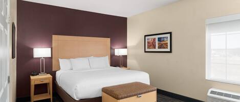 A hotel room with a large bed, two bedside tables with lamps, a small wooden bench, a framed picture on the wall, and a window with blinds.