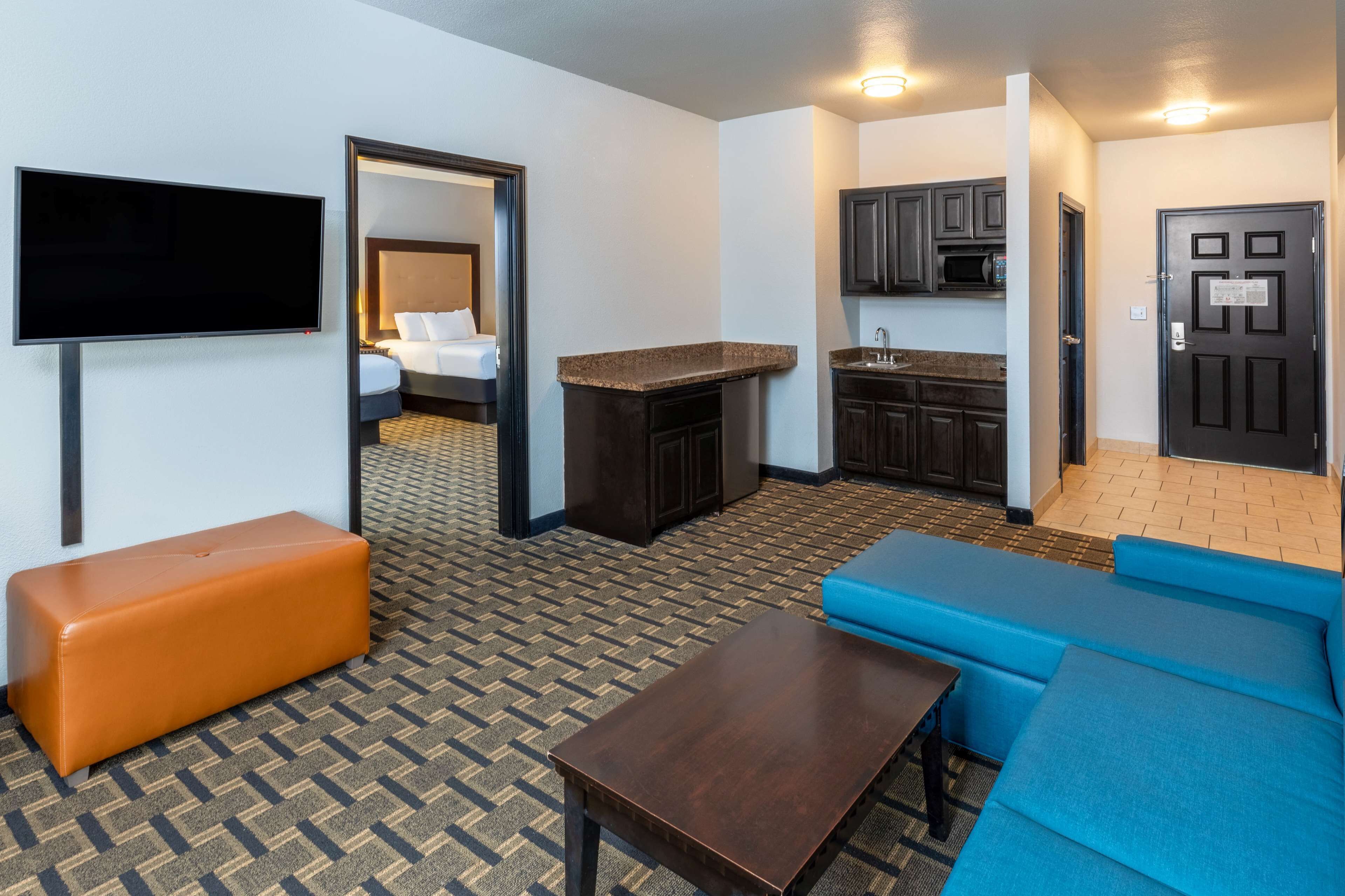 A hotel room with a kitchenette, a flat-screen TV, a sofa, a table, and a bench.
