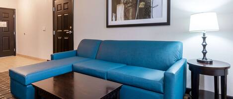 A living room with a blue sectional sofa, a dark wooden coffee table, a framed abstract artwork on the wall, and a small round table with a lamp.
