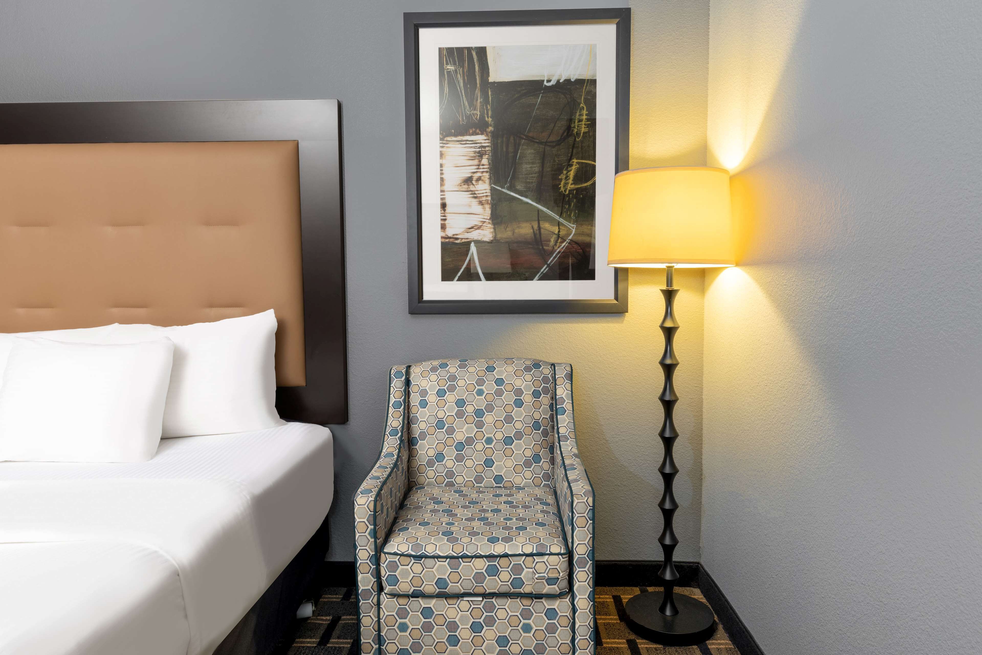 Une chambre d’hôtel avec un lit, un fauteuil à motifs, une lampe sur pied et une œuvre d’art encadrée accrochée au mur.