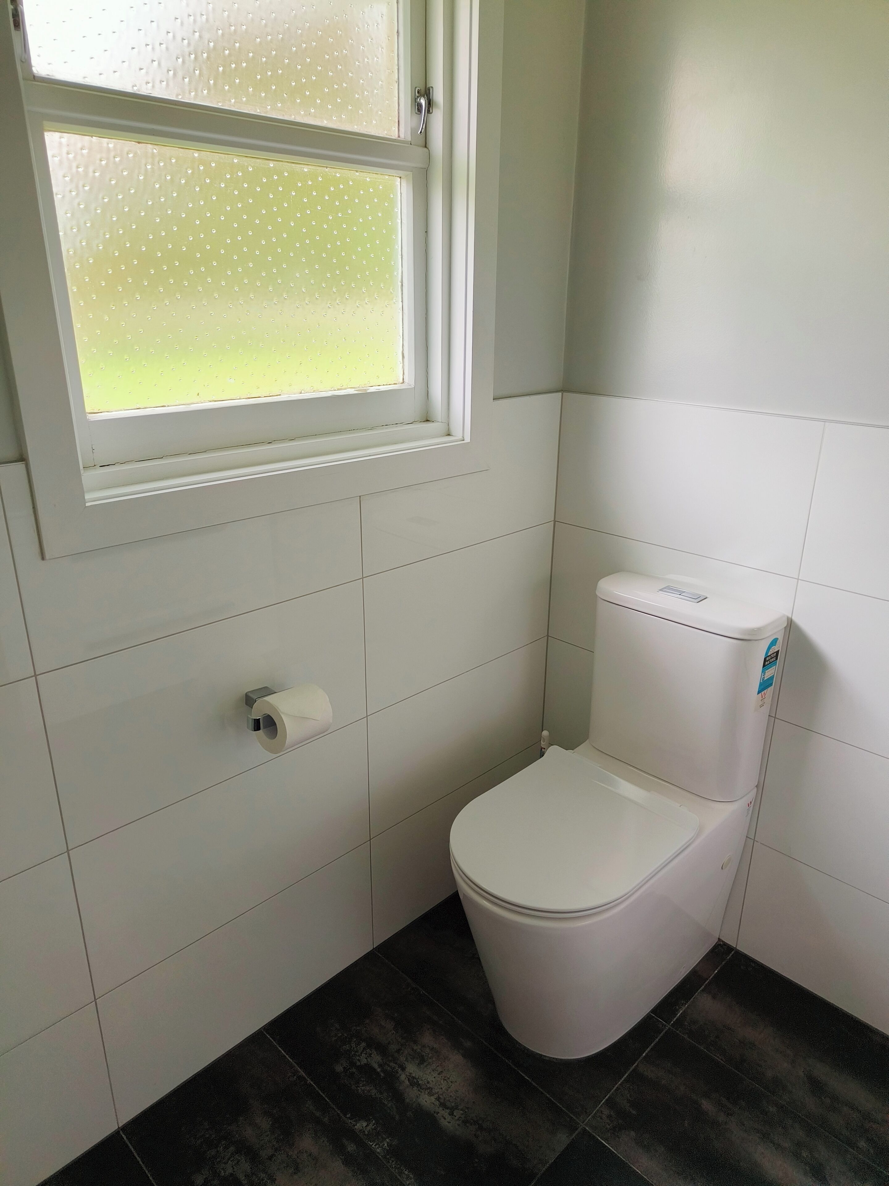Een kleine, schone badkamer met toilet, toiletrolhouder en een raam met gezandstraald glas.