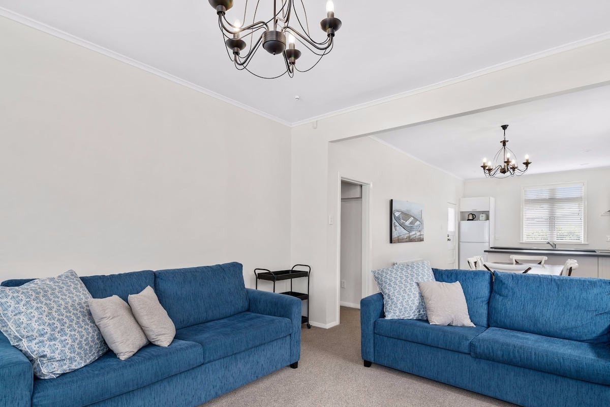A modern living room with two blue sofas, a chandelier, and a kitchen in the background.