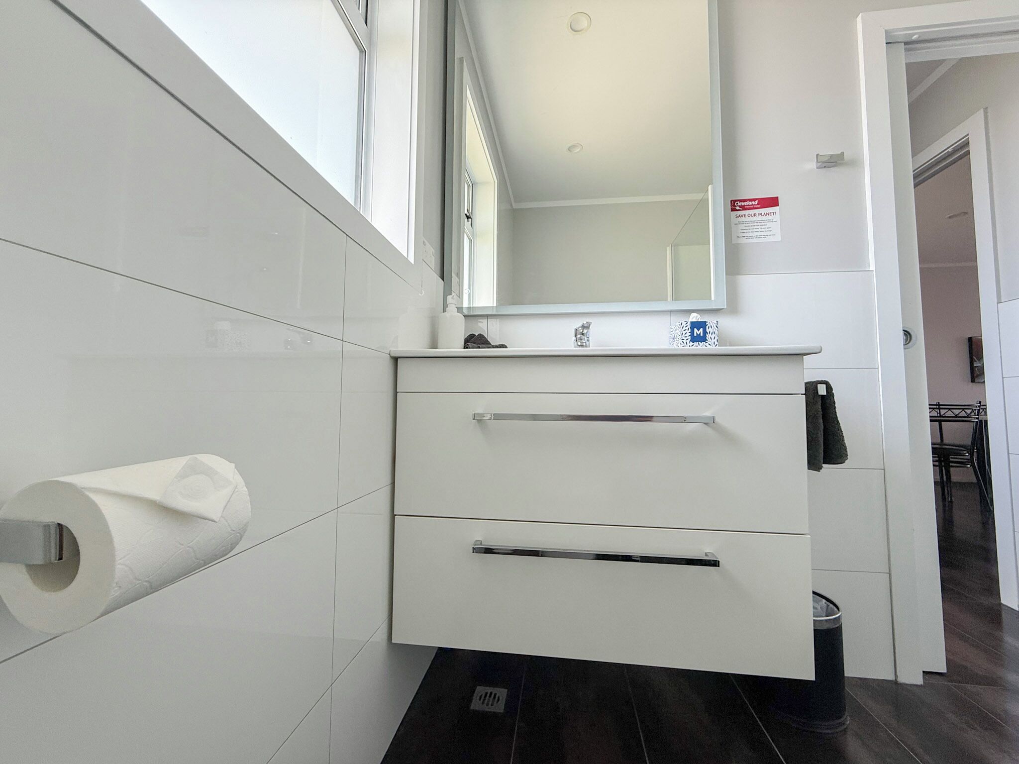 Un baño moderno con azulejos blancos, un dispensador de papel higiénico, un espejo y un lavamanos con grifo.