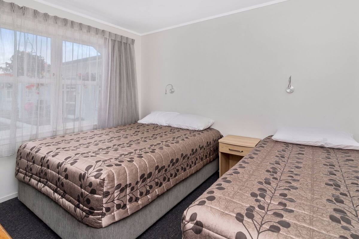 A hotel room with two beds, a window with sheer curtains, and a small bedside table.
