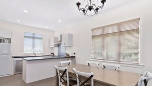 A modern kitchen with white cabinetry, a built-in island, and a dining area with a wooden table and white chairs. - ASURE Cleveland Rotorua (Rotorua)