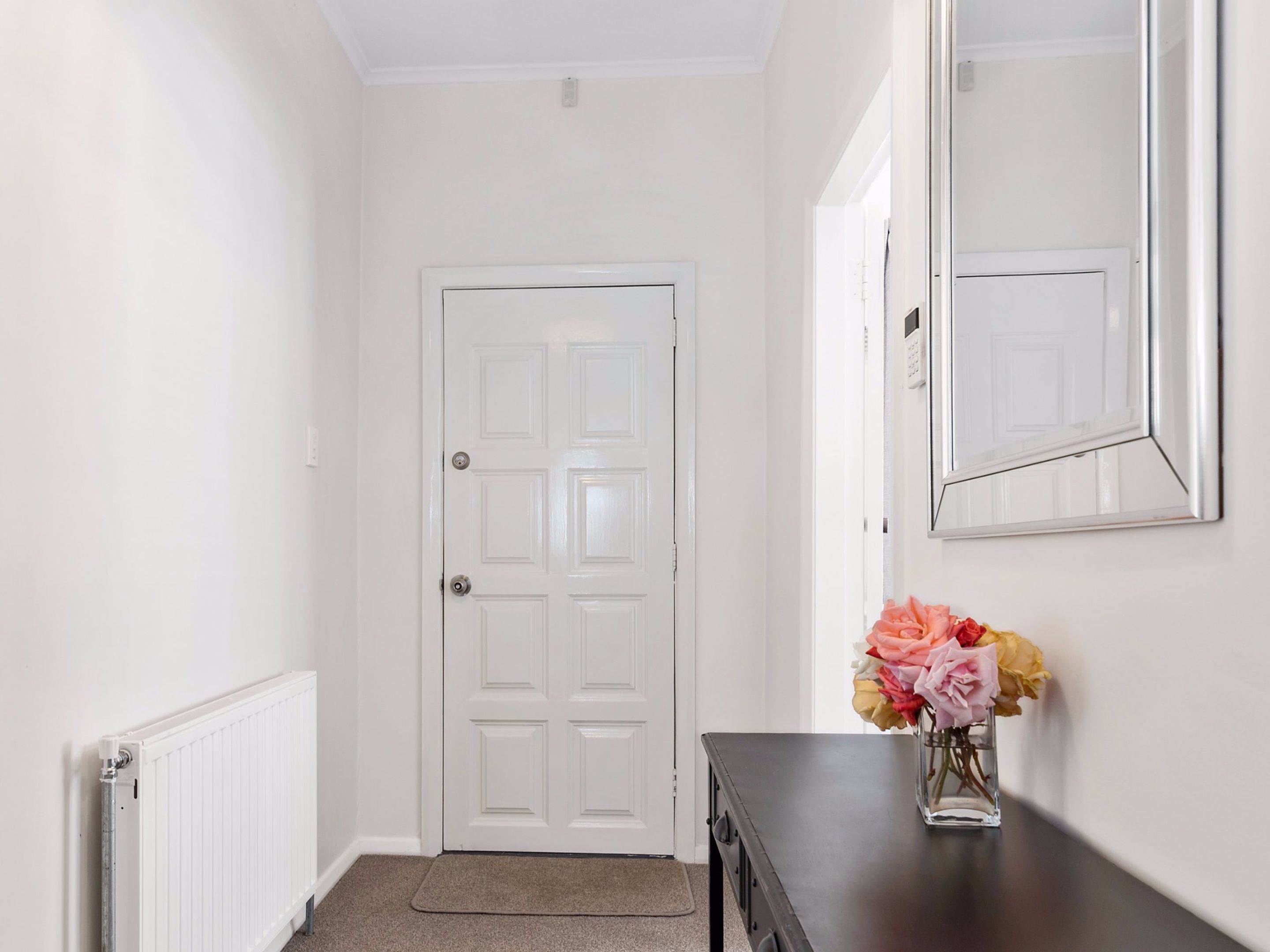 A hallway with a white door, a radiator, a dark wooden dresser, and a vase of flowers.