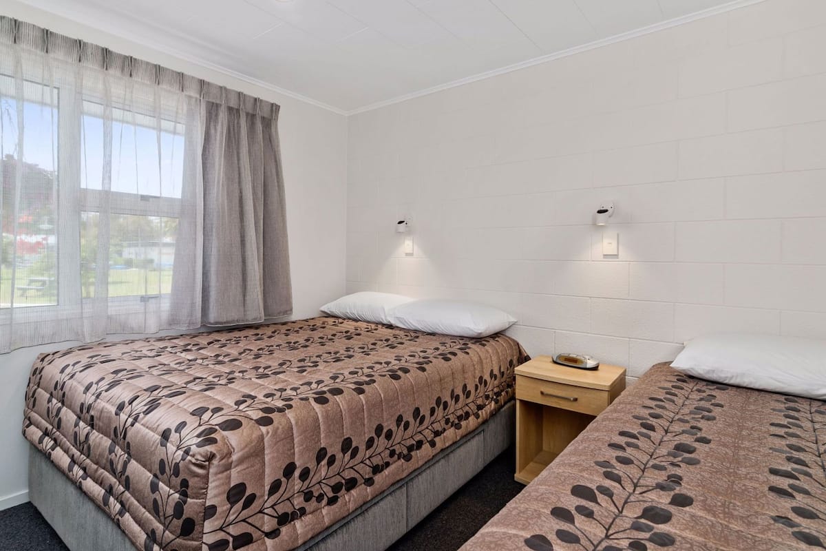 A bedroom with two beds, a window with curtains, a bedside table with a lamp, and a wall-mounted light.