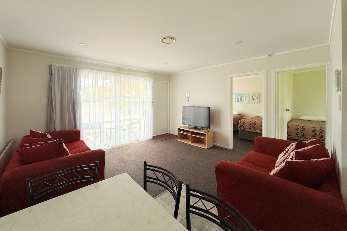 A living room with a TV, red sofas, a dining table, and a large window with curtains.