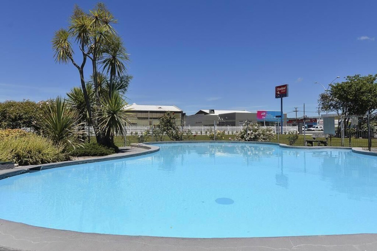 A swimming pool with a clear blue sky, surrounded by greenery and buildings in the background.