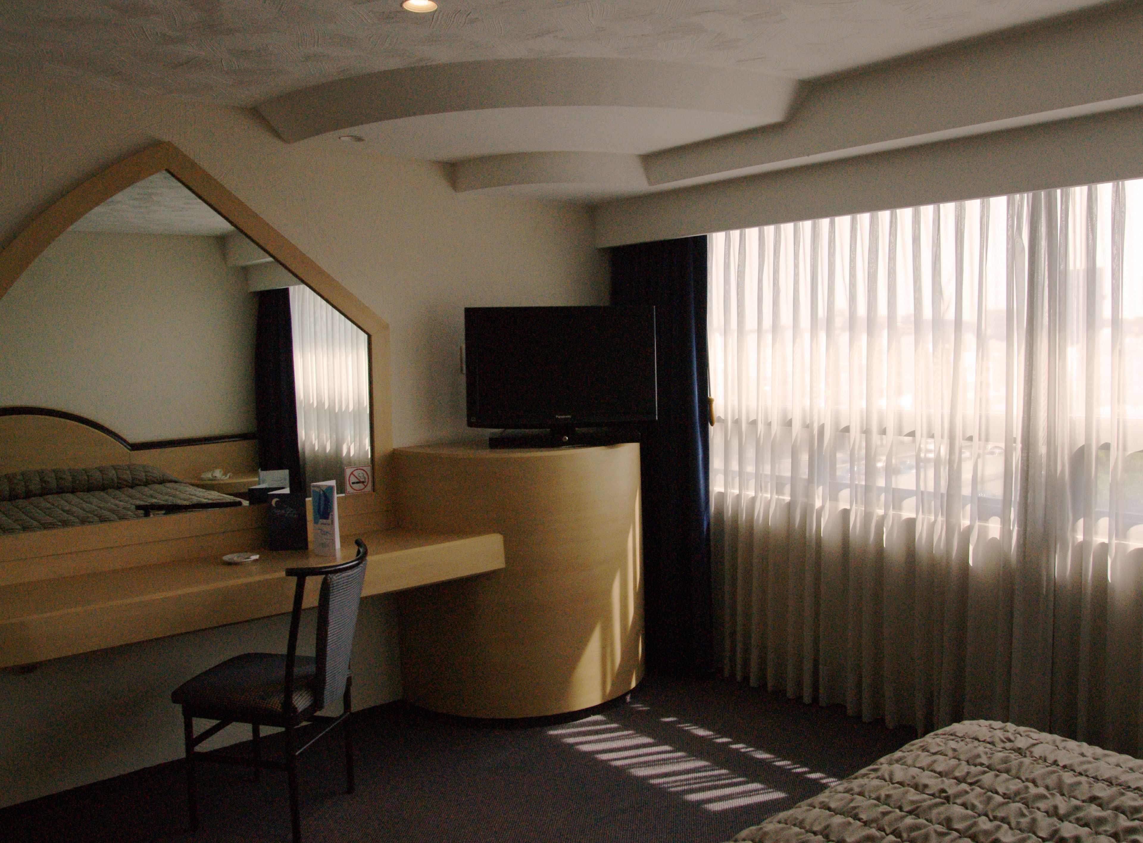 Habitació d'hotel amb un llit, una taula de treball amb una cadira, una televisió i una finestra gran amb cortines.