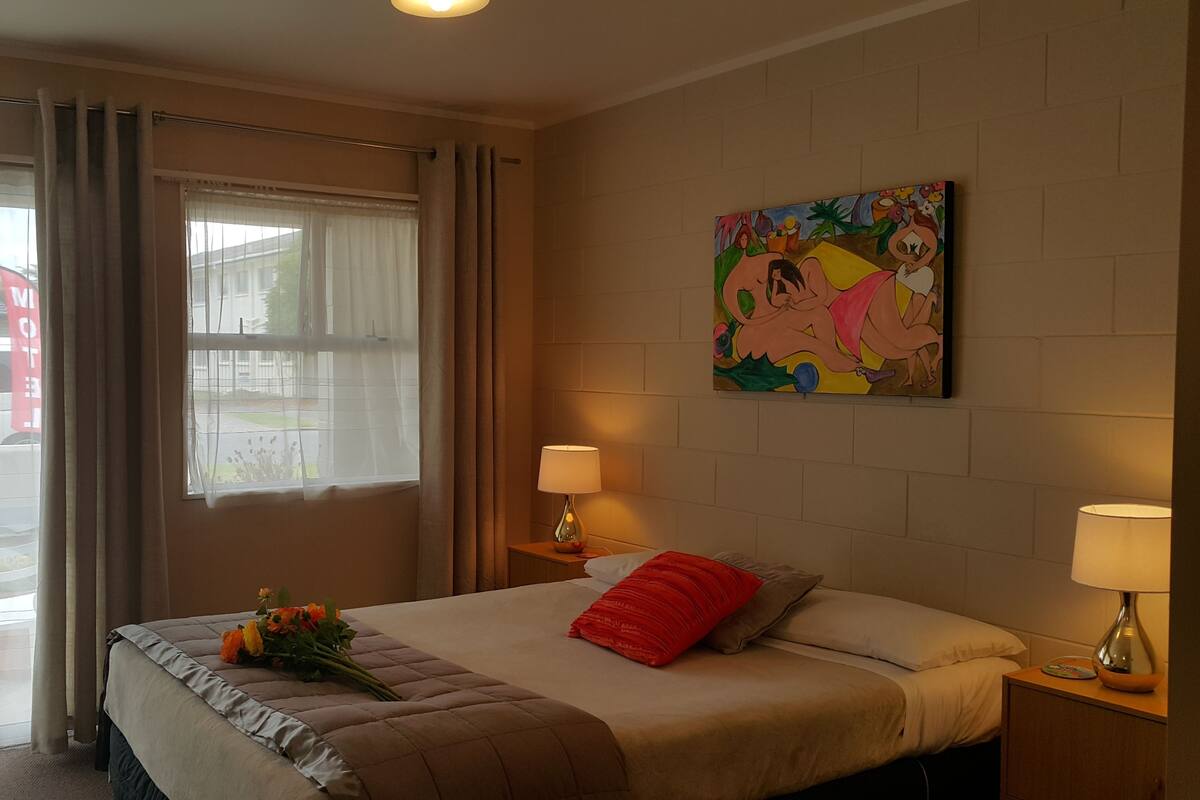 A hotel room with a bed, two bedside lamps, a window with curtains, a painting on the wall, and a bouquet of flowers on the bed.