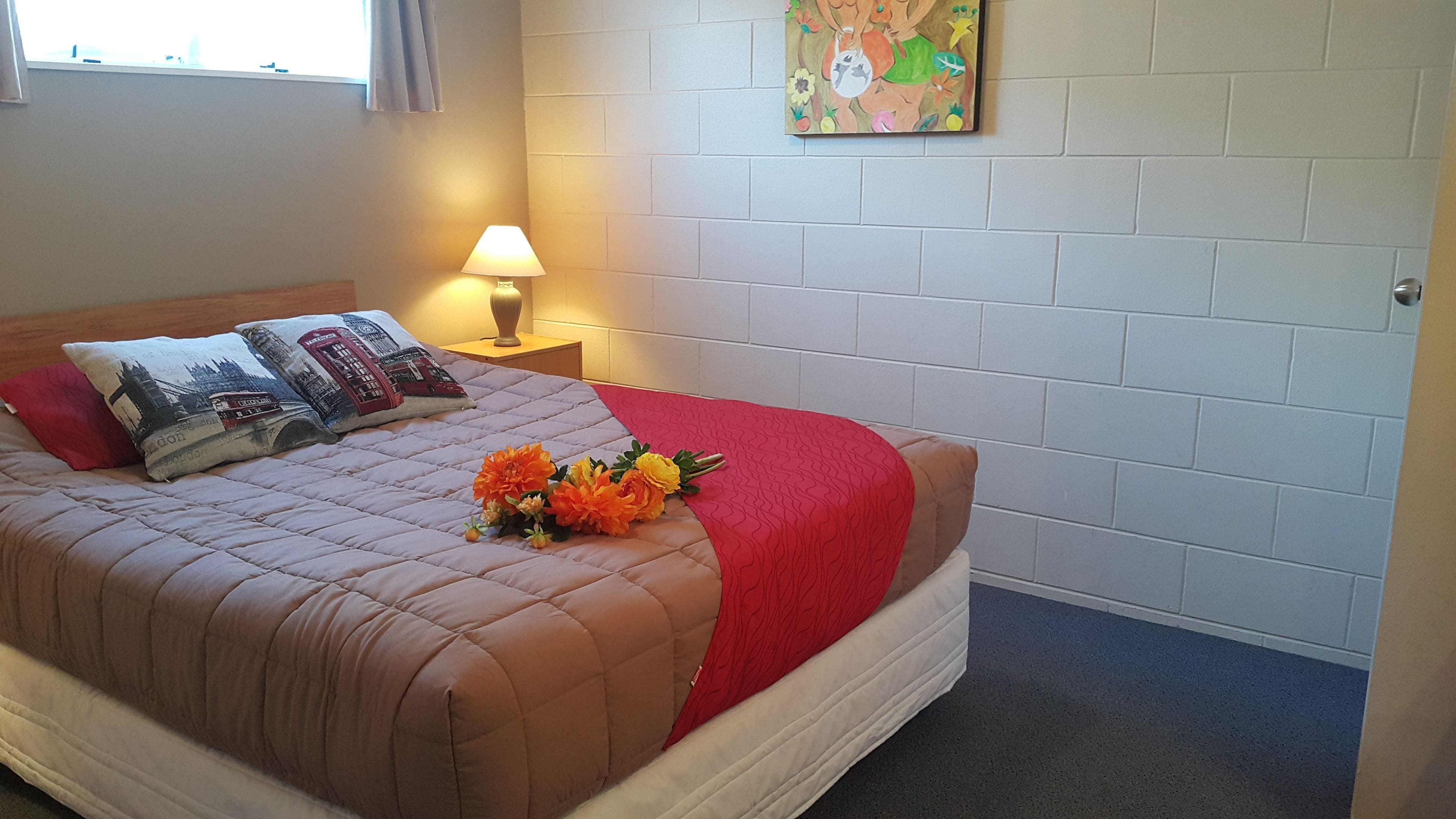 A neatly made bed with a red and brown quilt, two pillows with a London design, and a bouquet of flowers on the bed.