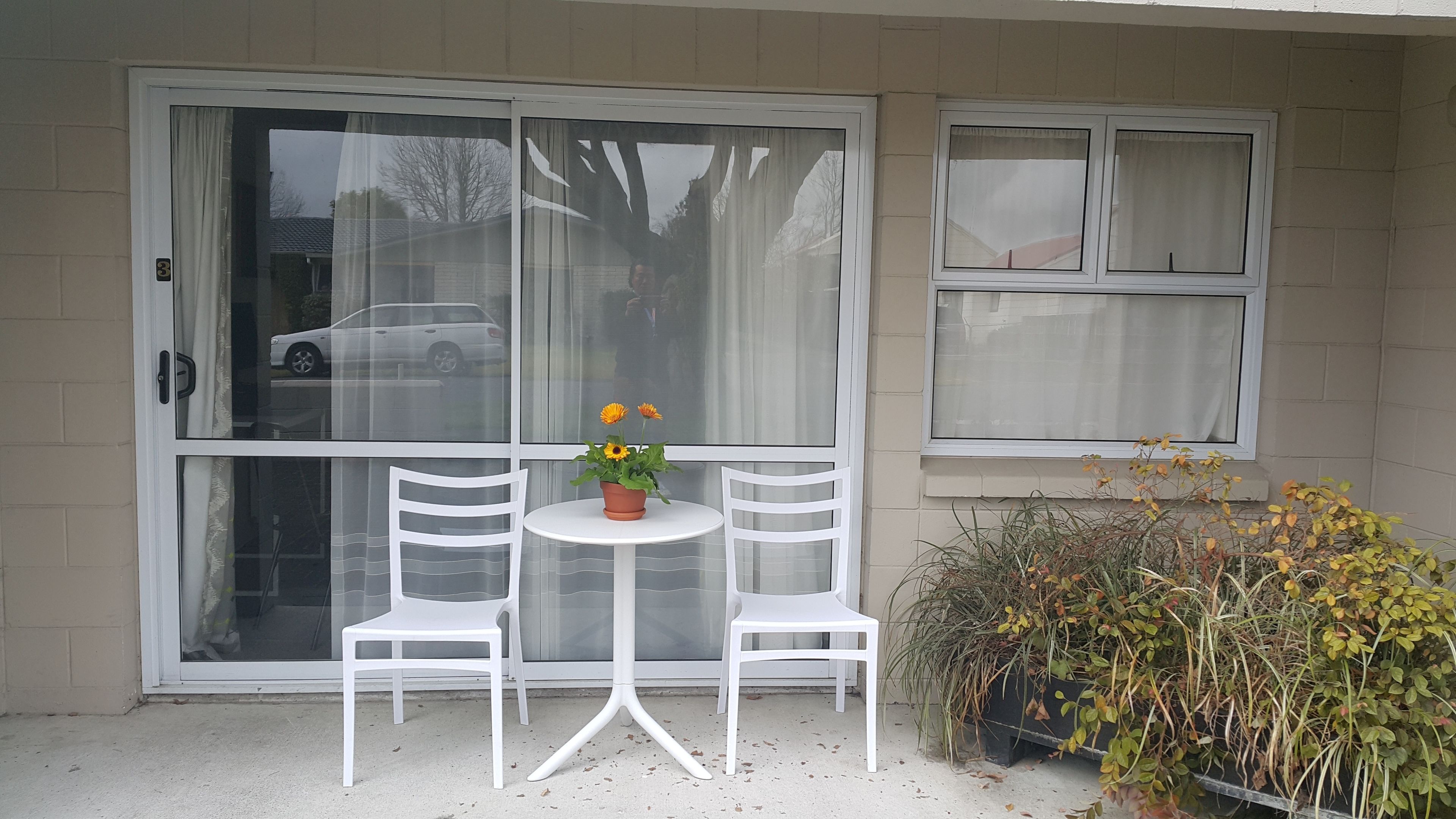 A small patio with two white chairs and a table, a potted plant with yellow flowers, and a window with a view of a car and trees.