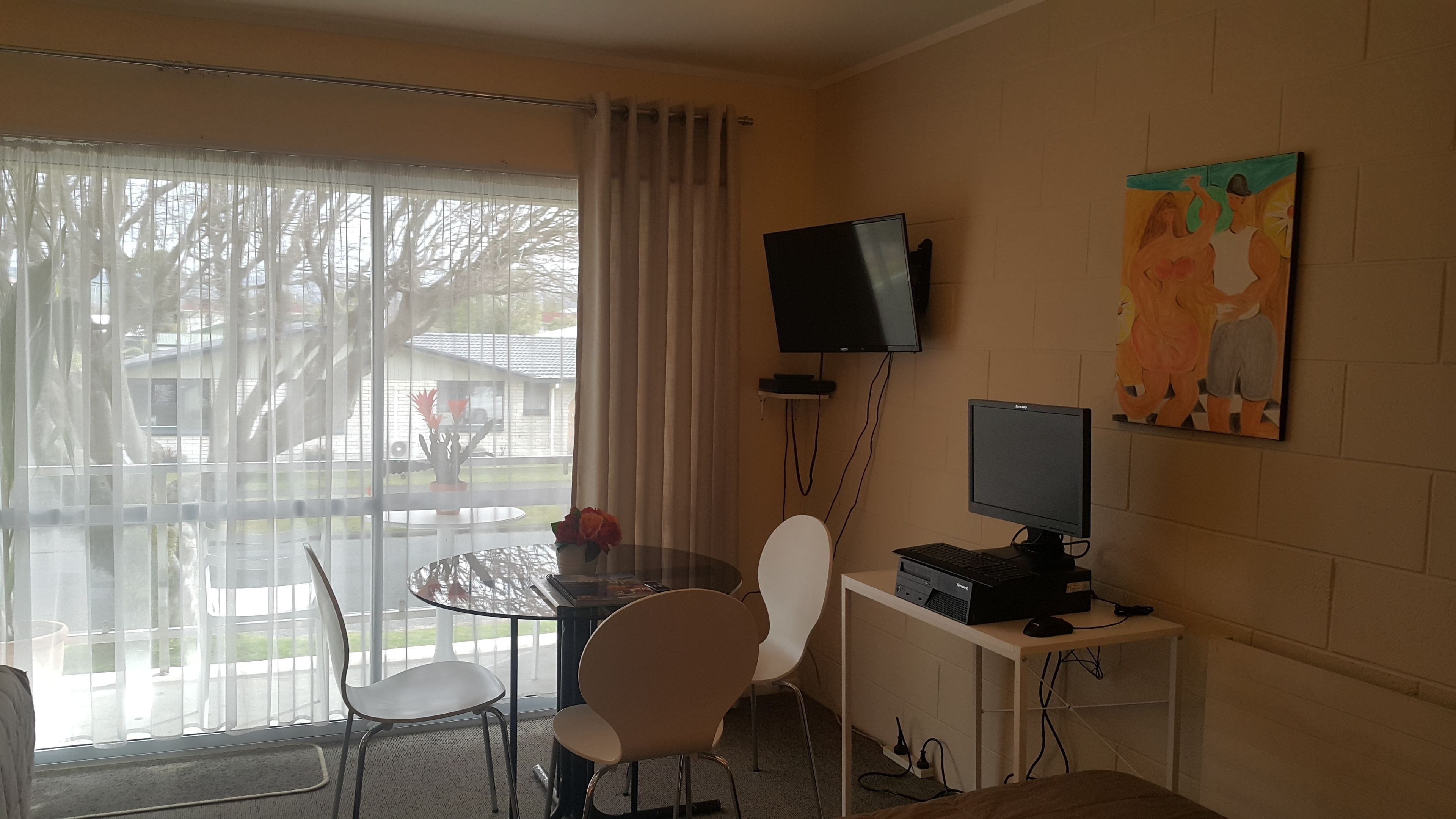 Una habitación con puerta de vidrio que da a un patio, una mesa redonda con sillas, un televisor de pantalla plana montado en la pared y un cuadro sobre el televisor.