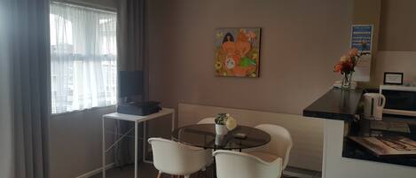Een knus verblijf met een eettafel, twee stoelen, een kitchenette met waterkoker en een schilderij aan de muur.