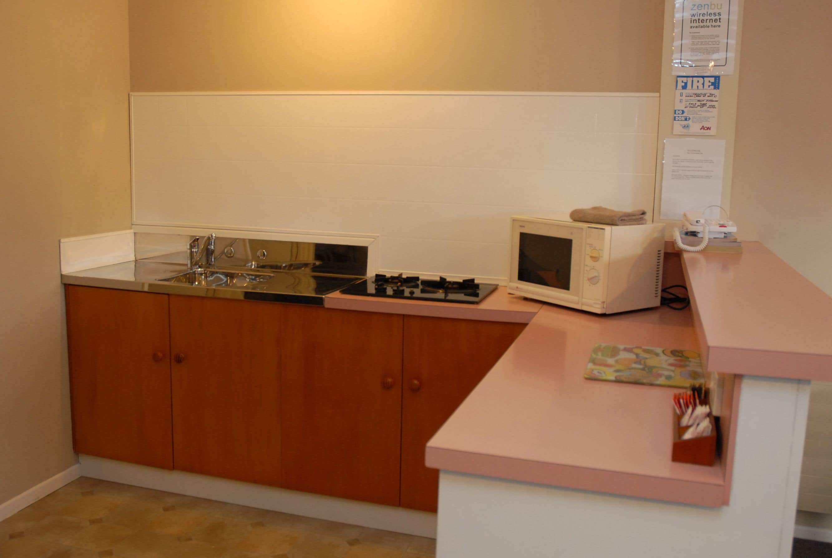 A compact kitchen with wooden cabinets, a sink, a microwave, and a small countertop with a telephone and a box of utensils.