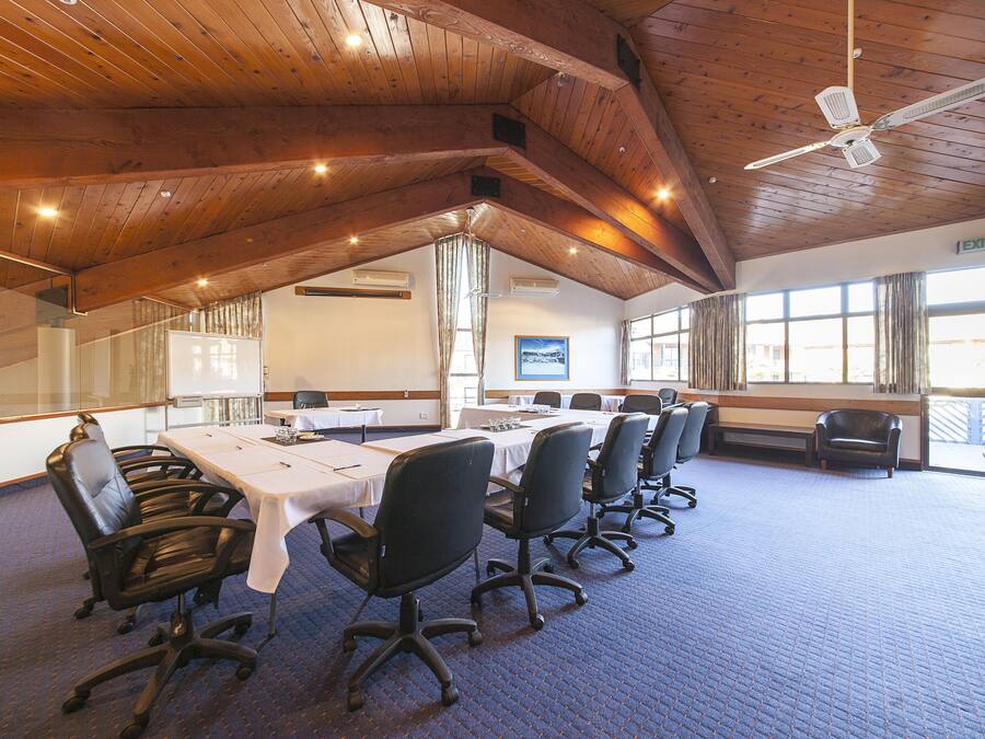 Un salón de conferencias con una larga mesa, sillas negras, vigas de madera en el techo y ventanas amplias.