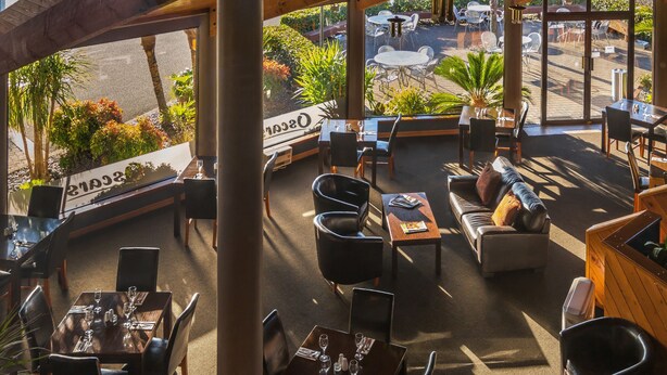 Un área de comedor al aire libre con mesas y sillas, un banco de madera y vistas a la calle y la vegetación.