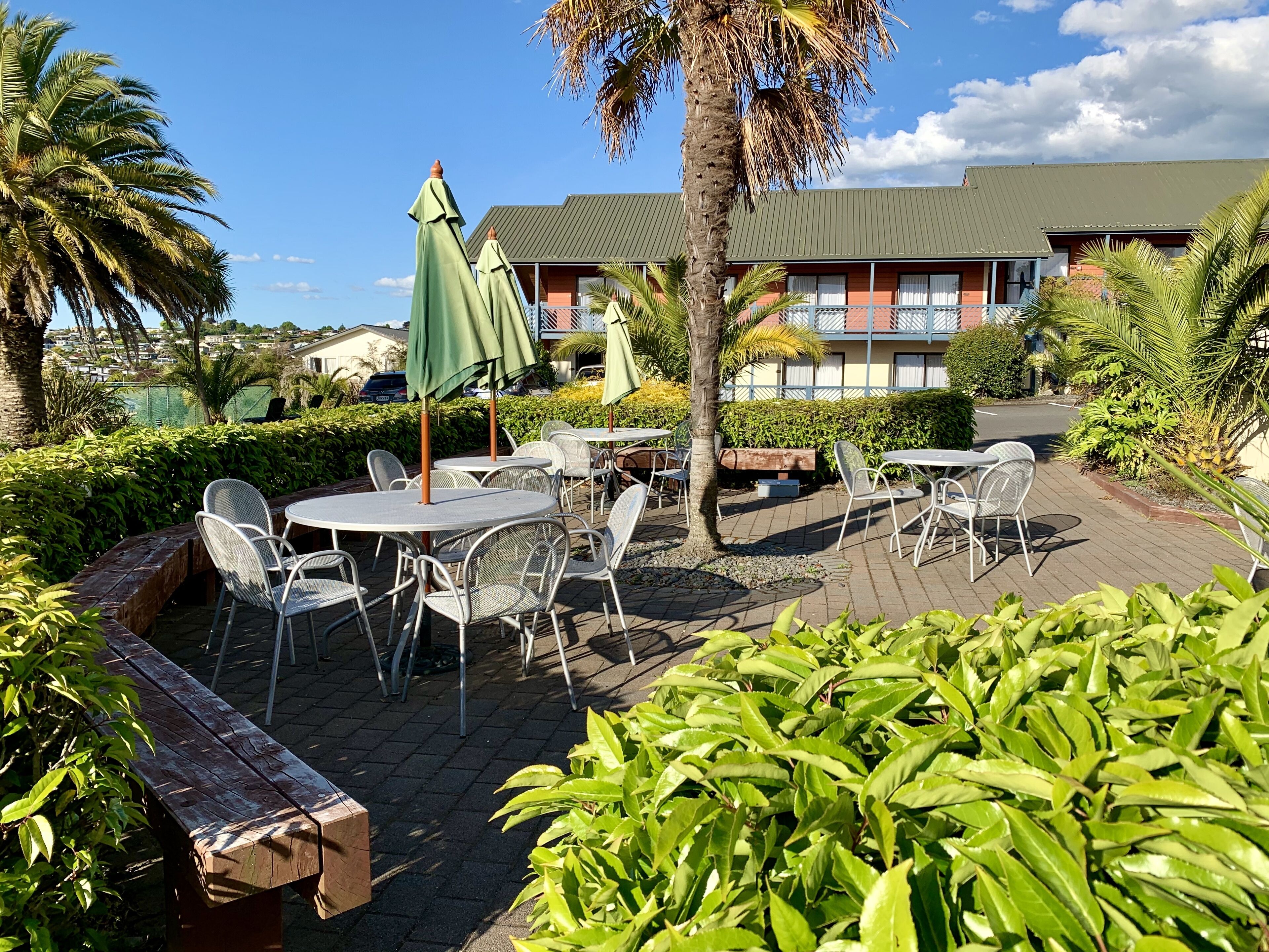 An outdoor patio area with round tables and chairs, surrounded by greenery and palm trees.