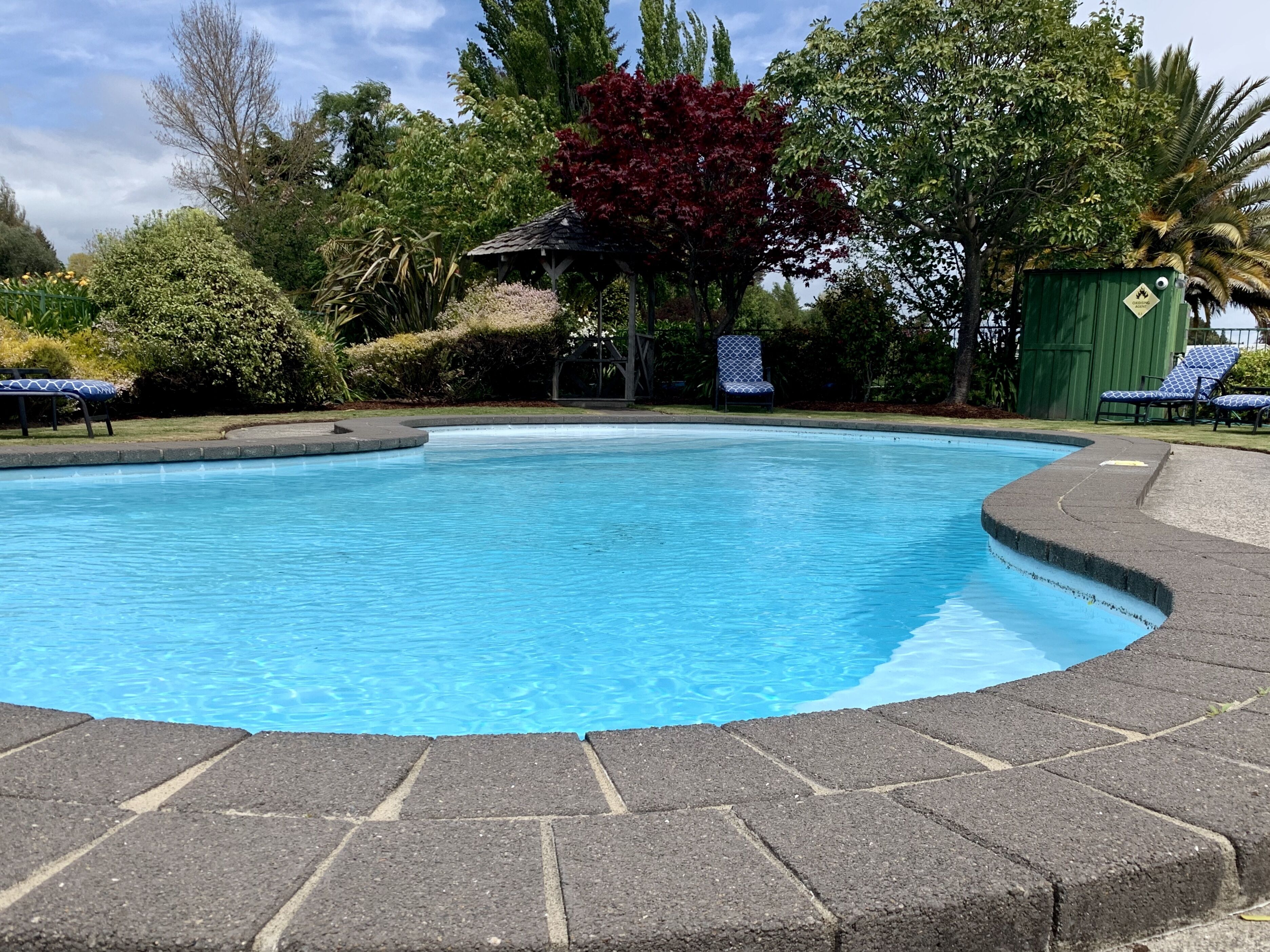 Uma piscina com área pavimentada ao redor, espreguiçadeiras e um gazebo.