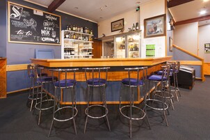 Una zona de bar con mostrador de madera, taburetes metálicos con cojines azules y una pizarra con la carta de bebidas y ofertas.