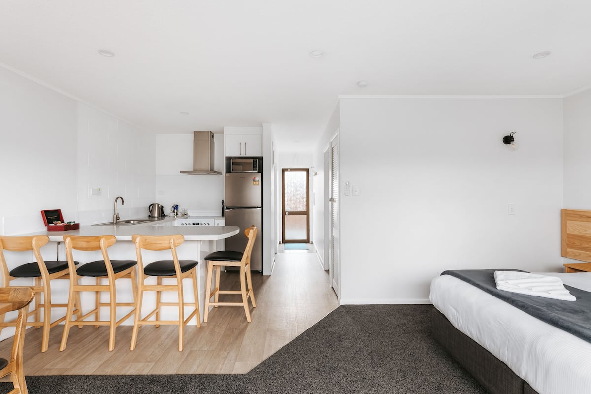 Ein modernes, minimalistisches Hotelzimmer mit Küchenzeile, einem Esstisch mit Stühlen und einem Bett mit dunklem Kopfteil.