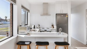 A modern kitchen with white cabinets, a stainless steel refrigerator, a microwave, and a gas stove. There are three black stools at the counter. - Cobblestone Court Motel – Wenzel Motels (Tauranga)