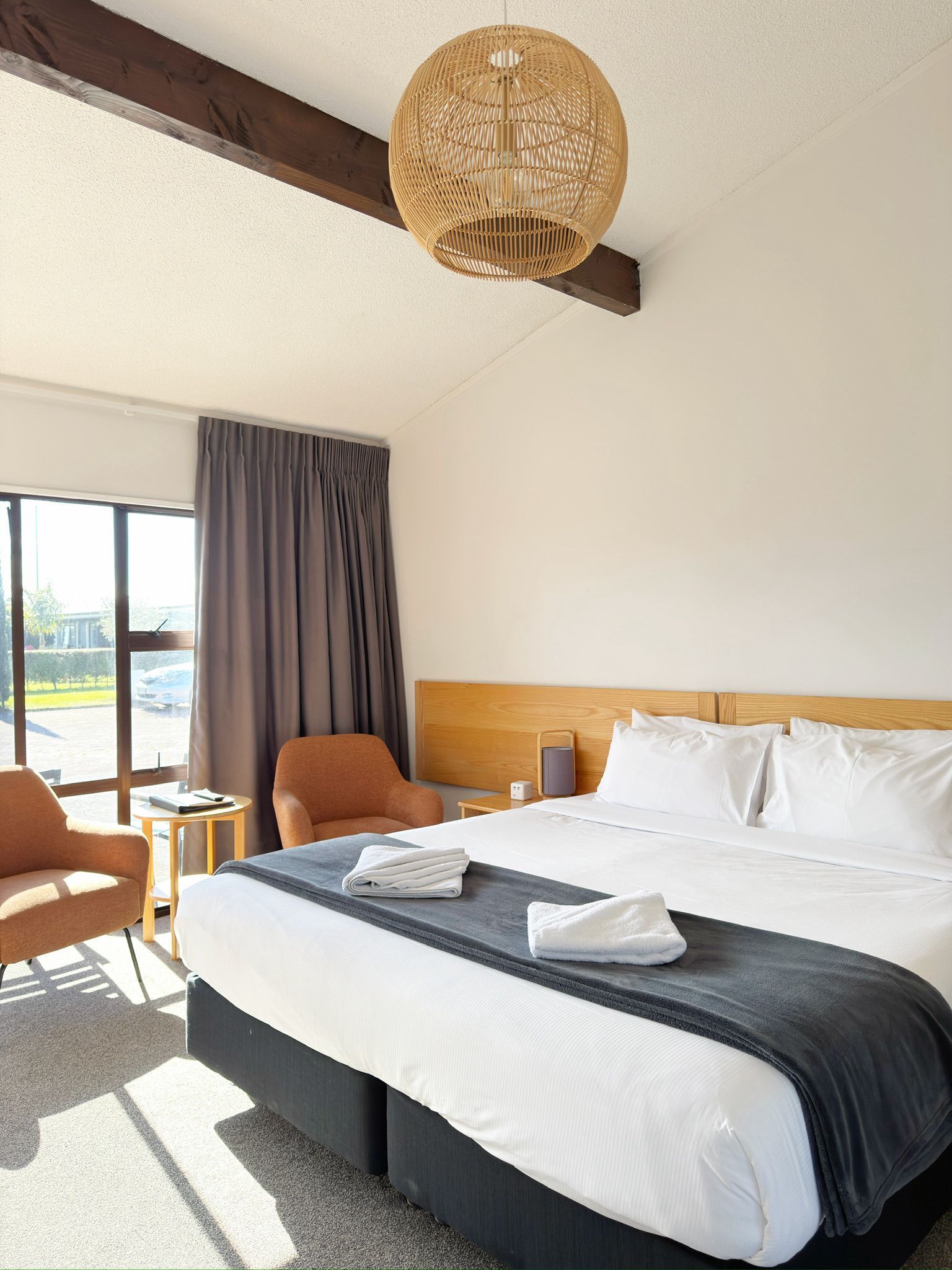 A hotel room with a large bed, a wooden headboard, a wicker ceiling light, a window with curtains, and a view of the outside.