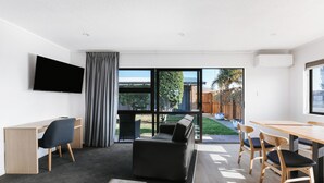 A modern living room with a flat-screen TV, a sofa, a dining table with chairs, and large windows offering a view of the outdoors. - Cobblestone Court Motel – Wenzel Motels (Tauranga)