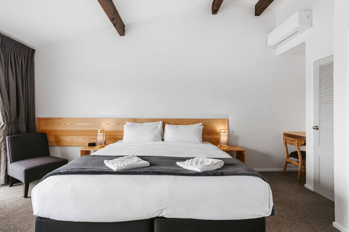 Ein Hotelzimmer mit Holz-Kopfteil, einem Bett mit weißer und grauer Bettwäsche, einem Sessel, einem Schreibtisch und einer Klimaanlage.