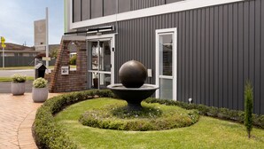 A modern building with a landscaped garden featuring a spherical fountain, a key mailbox, and a brick archway. - Cobblestone Court Motel – Wenzel Motels (Tauranga)