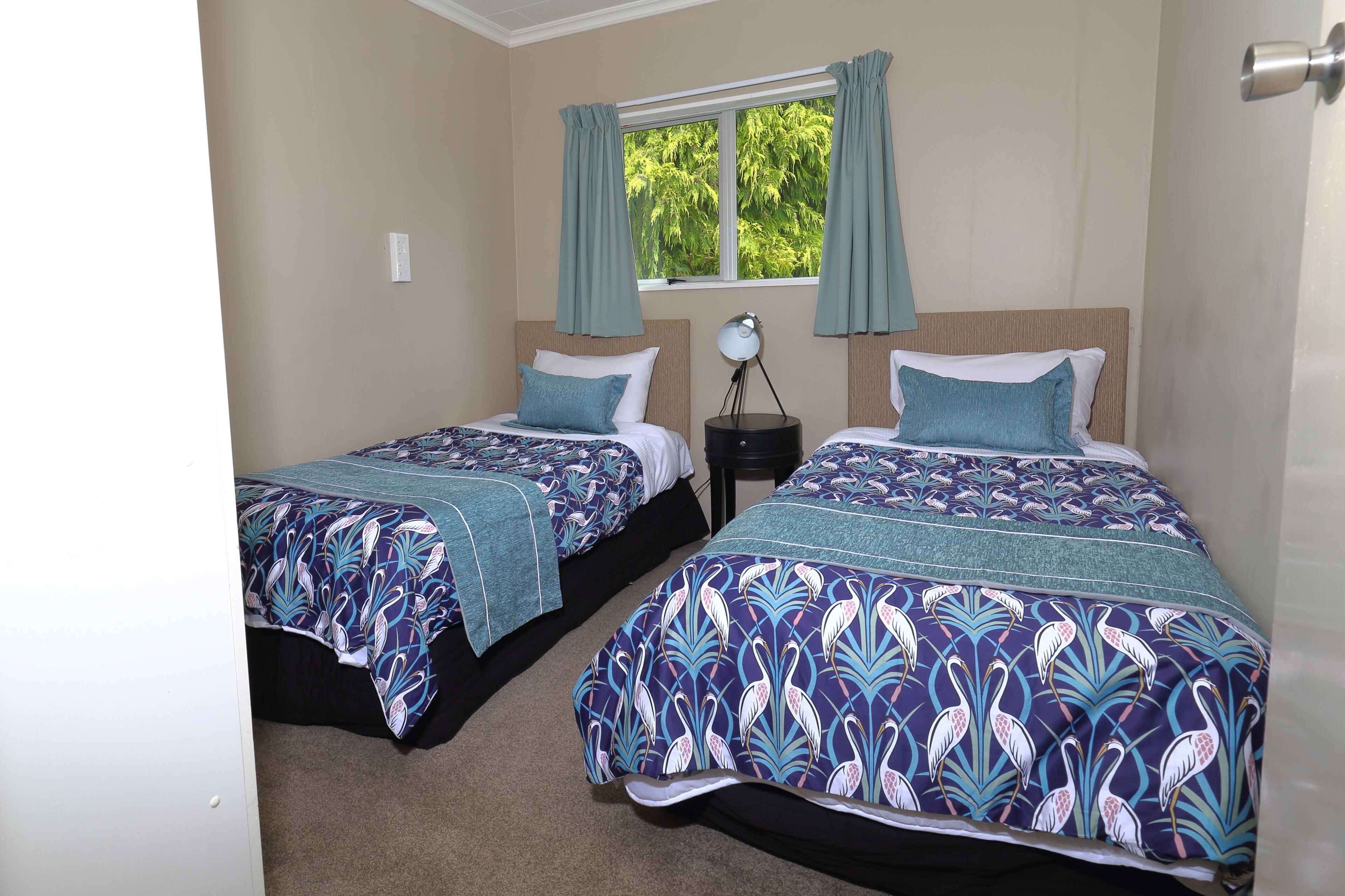 A bedroom with two beds, each with a patterned bedspread, a nightstand with a lamp, and a window with curtains.