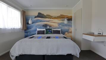 Un dormitorio con una cama, un mural de montañas y nubes, una mesita de noche con una lámpara, y una estantería con un libro y un dispositivo.