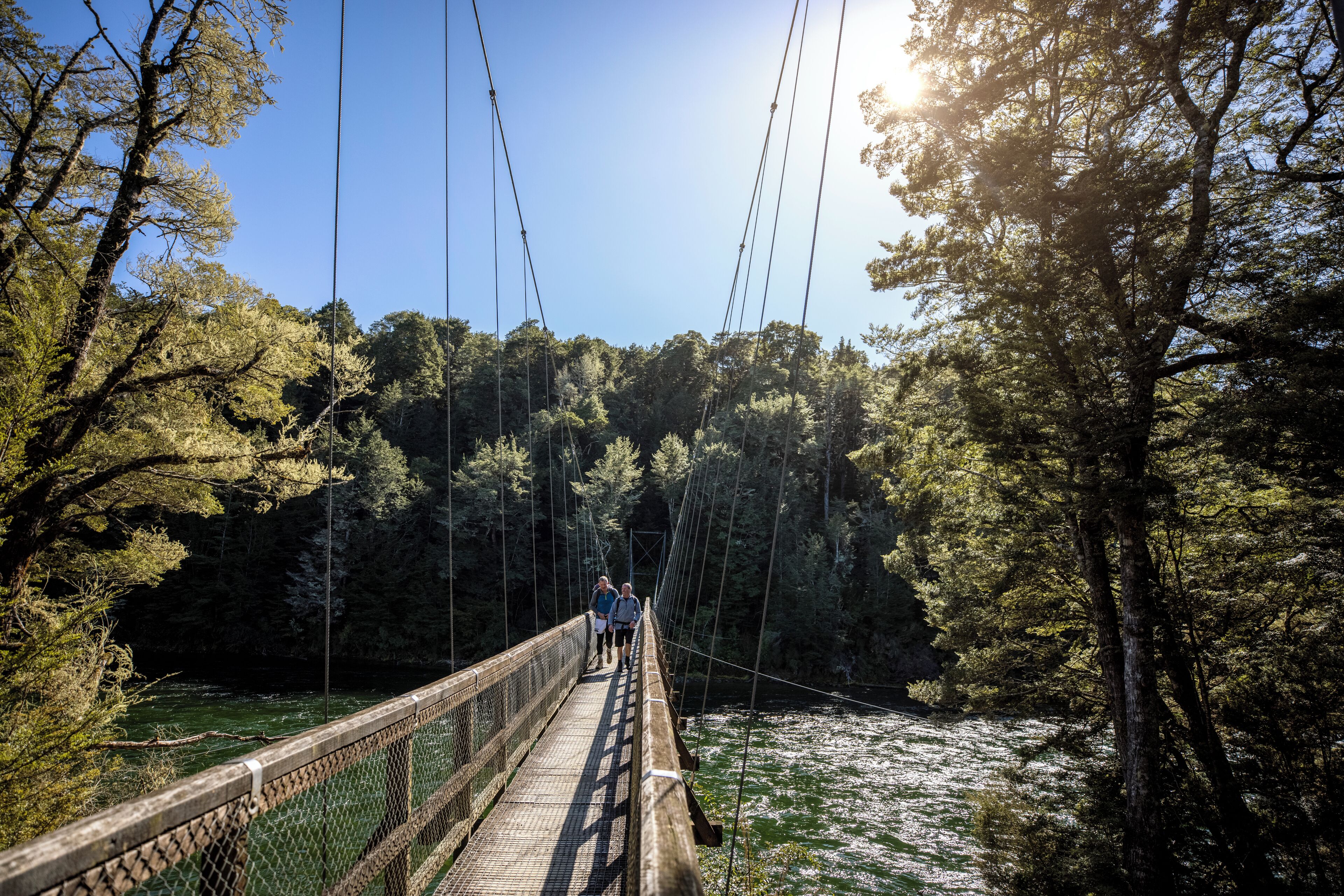 Eine Hängebrücke mit Holzbelag und Metallgeländern, umgeben von dichtem Wald und einem Fluss darunter.