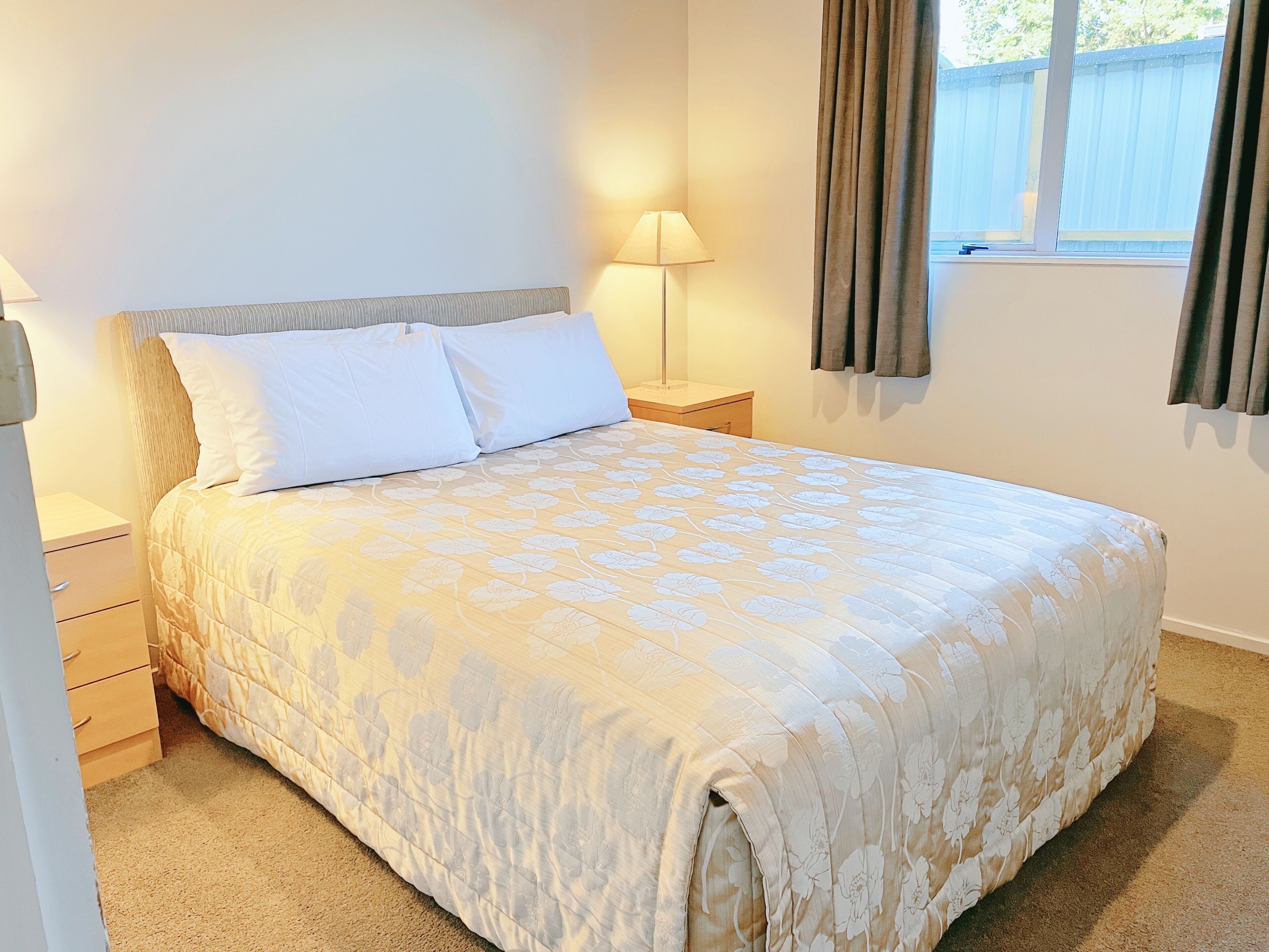 A neatly made bed with a floral-patterned bedspread, a bedside lamp, and a window with curtains.