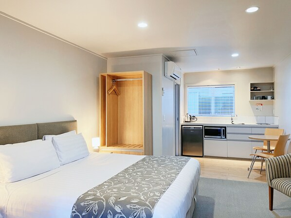 A hotel room with a bed, a wooden wardrobe, a kitchenette with a refrigerator, microwave, and a small dining table with chairs. - Anchorage Motel Apartments (Te Anau)
