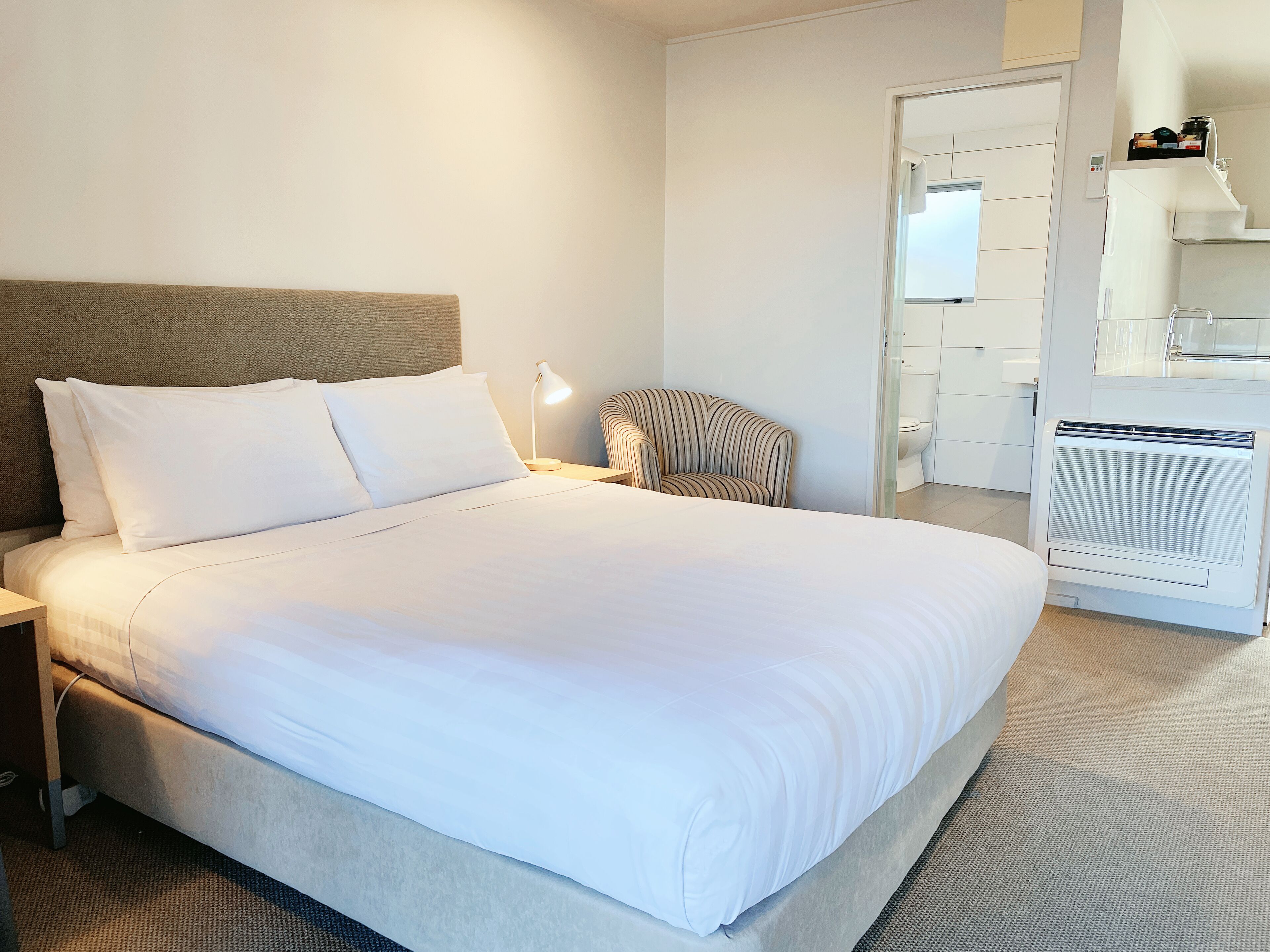 A neatly made bed with white linens, a bedside lamp, a striped chair, and a white radiator.