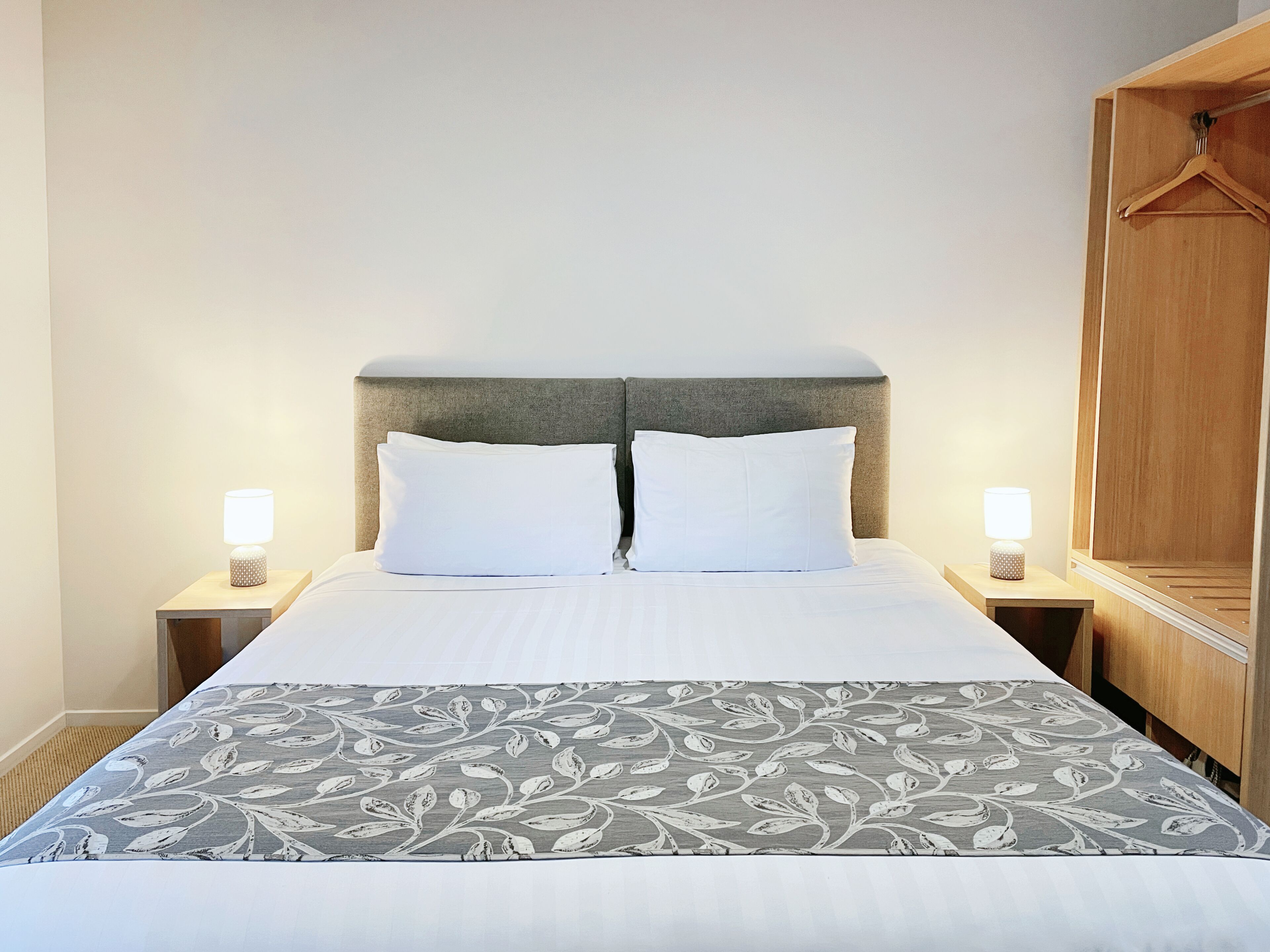 A neatly made bed with a grey and white patterned bedspread, two white pillows, and a grey headboard. There are two bedside tables with lamps on either side of the bed.
