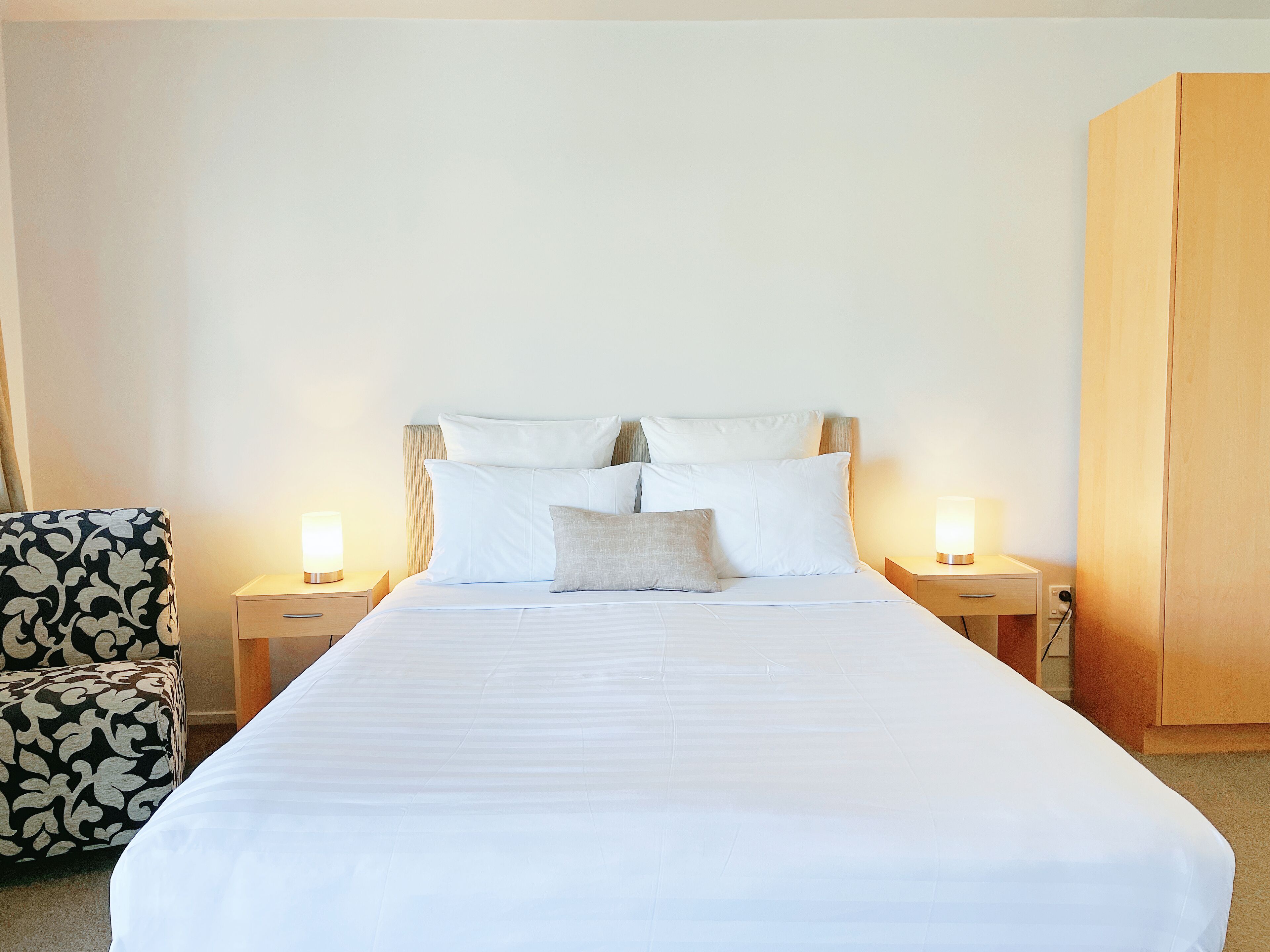 A neatly made bed with white linens and multiple pillows, flanked by two bedside tables with lamps, a patterned armchair, and a wooden wardrobe.