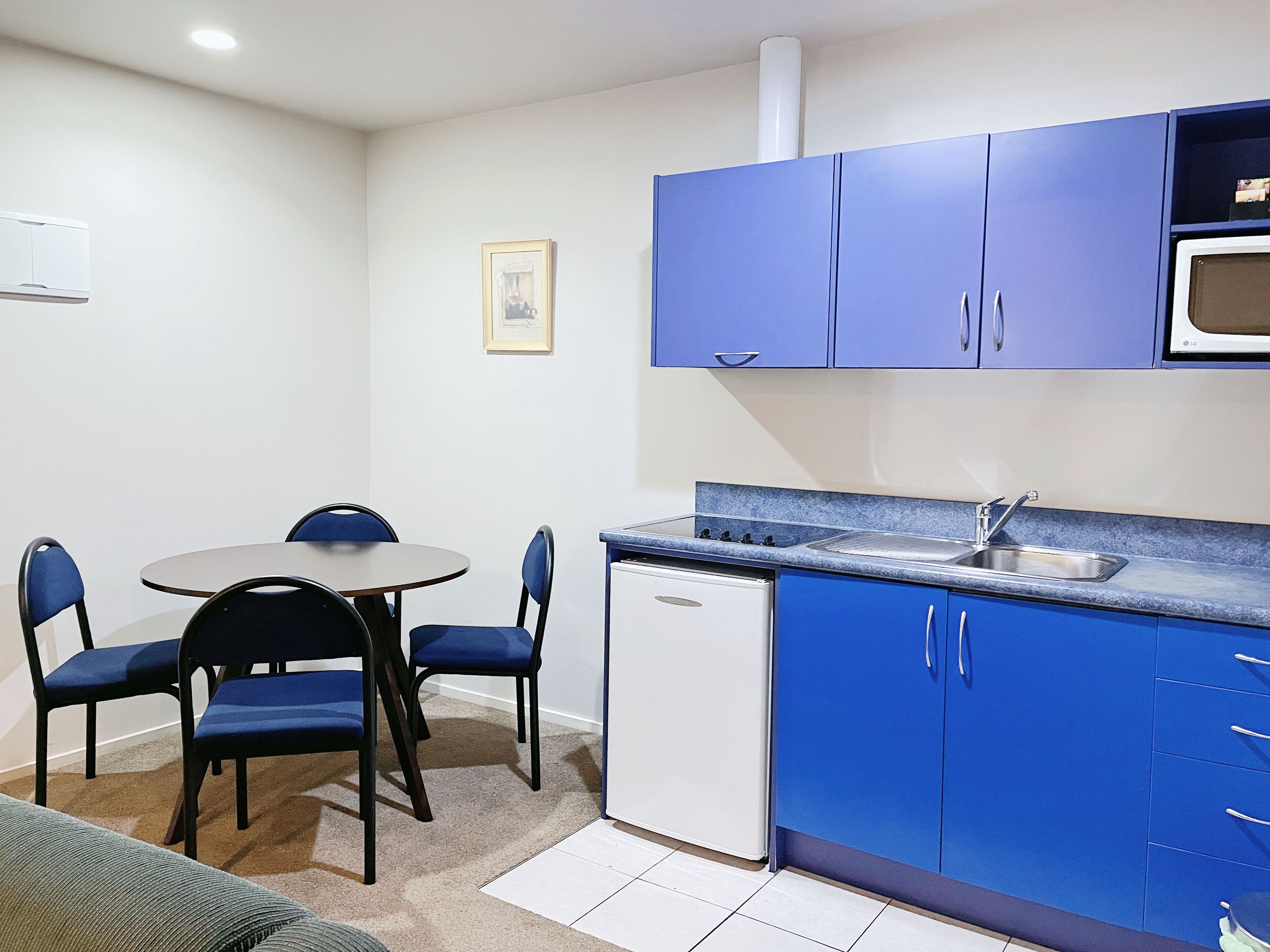 Una cocina compacta con gabinetes azules, un refrigerador blanco, un microondas y un lavamanos. Hay una pequeña mesa de comedor con cuatro sillas.