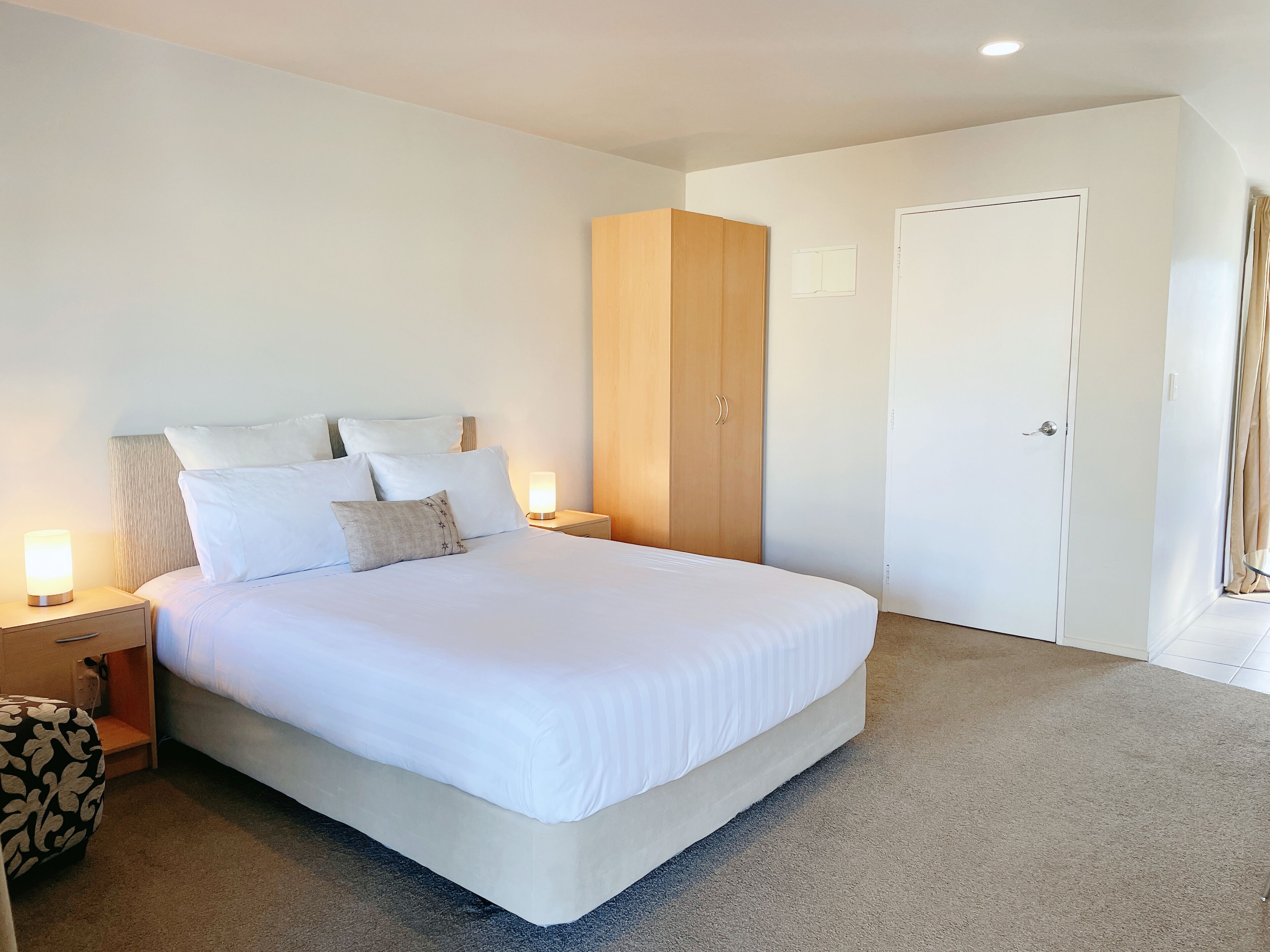 A neatly made bed with white linens, a wooden wardrobe, a bedside lamp, and a white door.