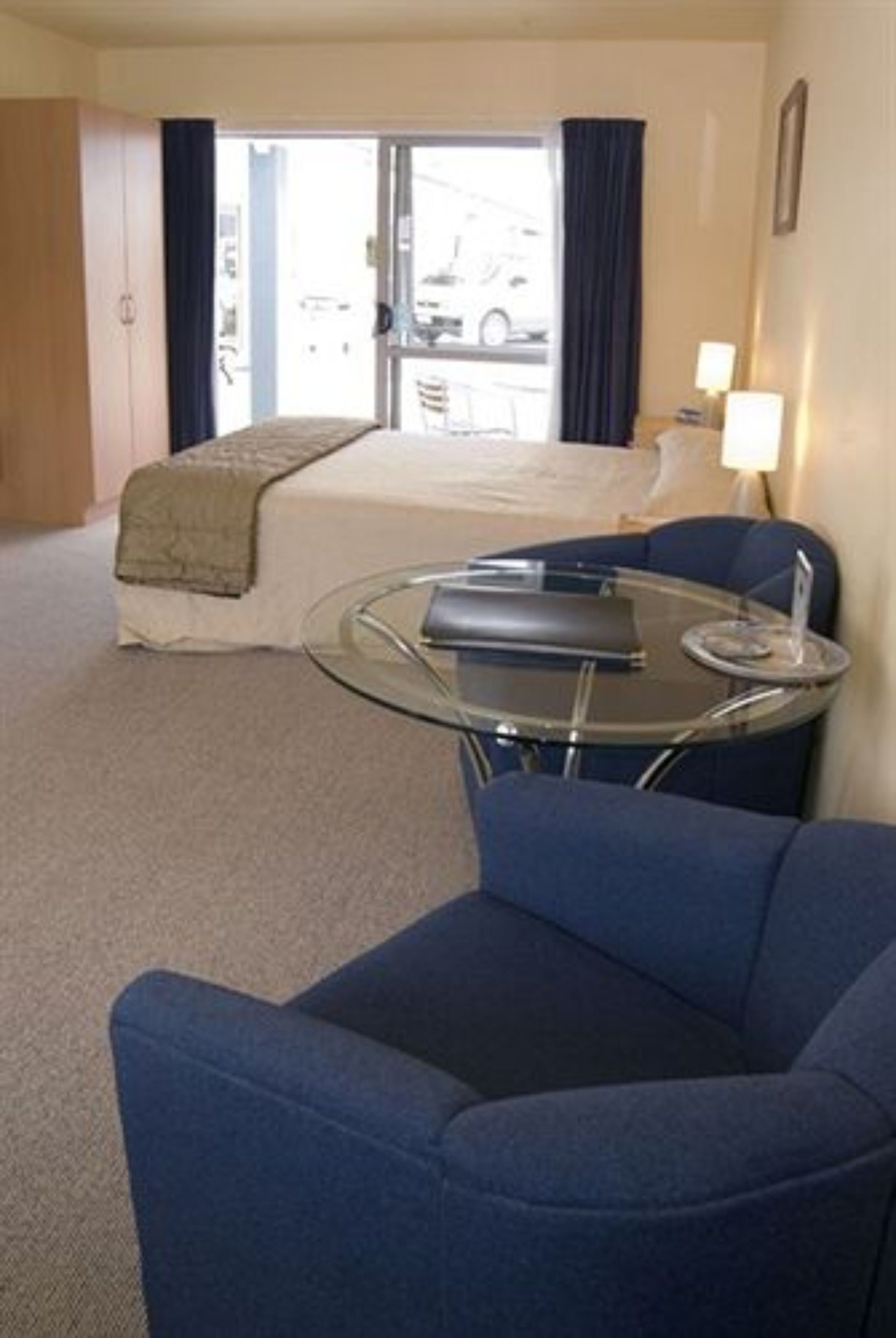 A hotel room with a bed, a glass table, a blue armchair, and a view of the street through the window.