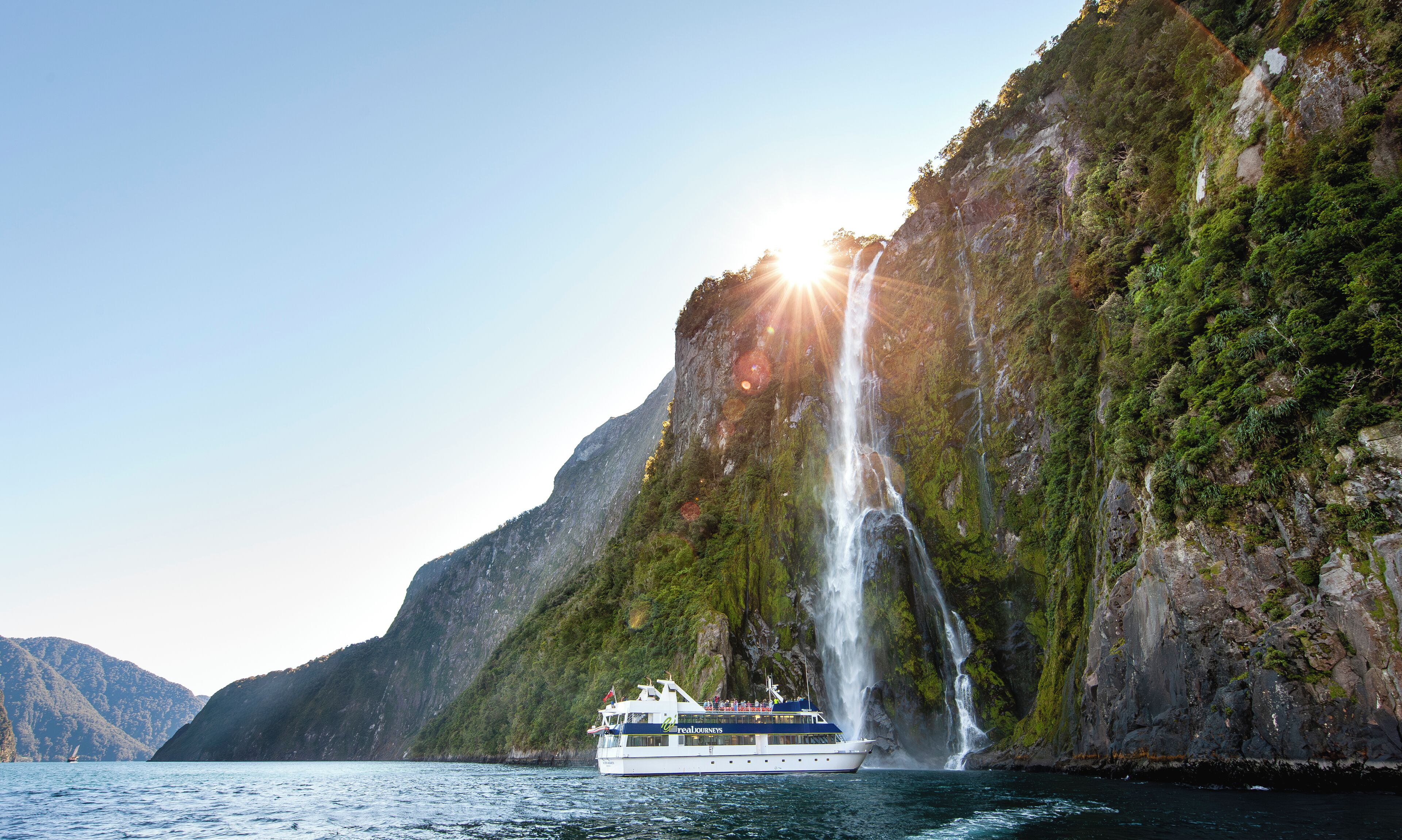 Un bote con la leyenda 'real journeys' en su lateral, ubicado cerca de una cascada y un acantilado escarpado.