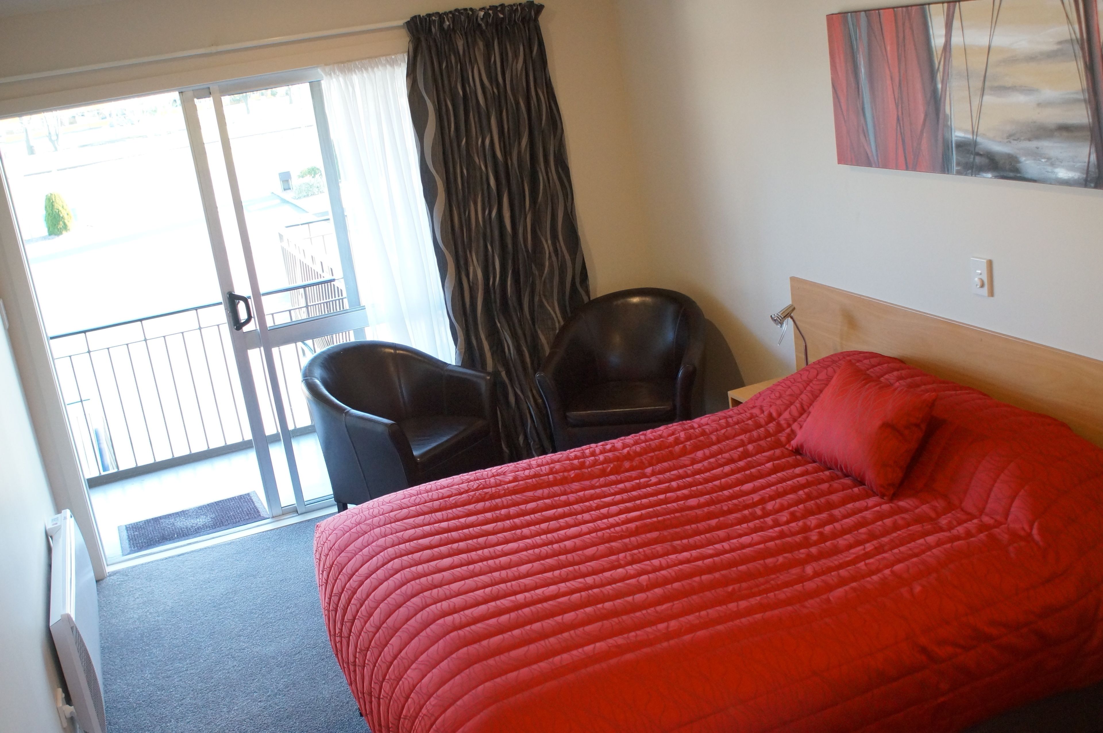 A hotel room with a red bed, a chair, a window with curtains, and a painting on the wall.