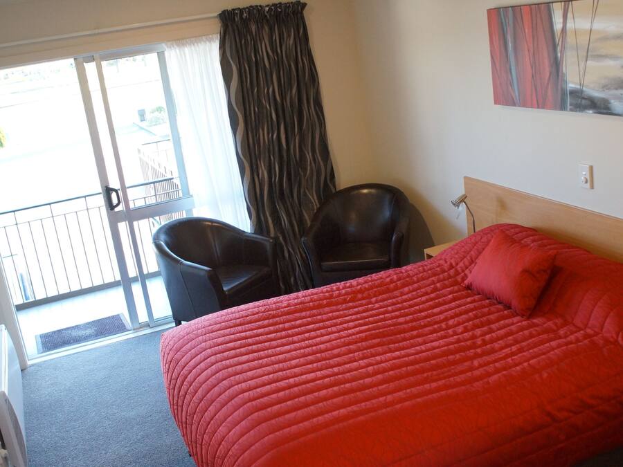 A hotel room with a red bed, a chair, a window with curtains, and a painting on the wall.