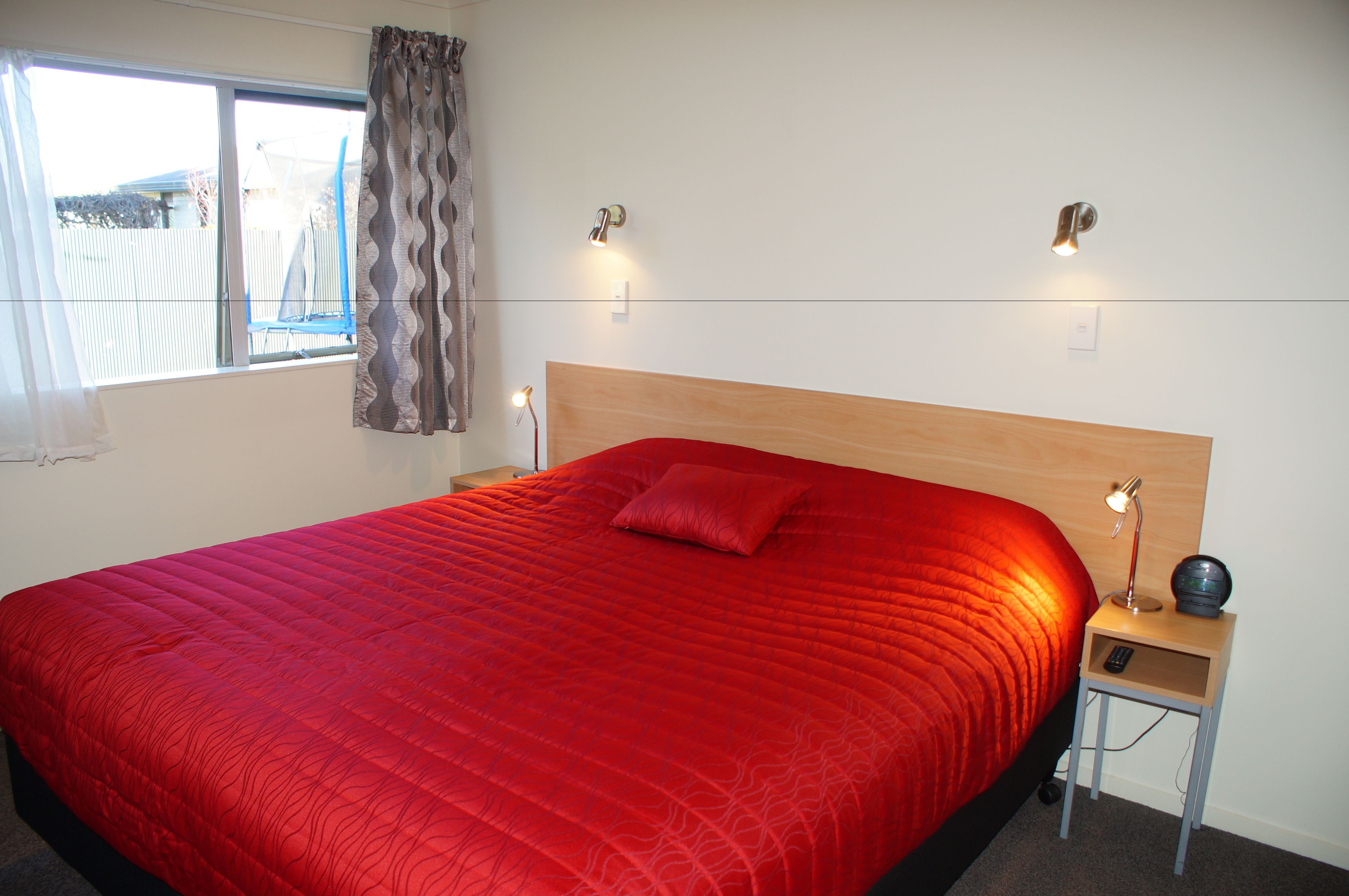 A neatly made bed with a red bedspread and a single pillow, a bedside table with a lamp, and a window with curtains.